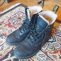 Front and top view of black leather Doc Martens boots with fur lining at the collar, showing lace-up design and yellow stitch details.