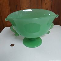 Green jadeite glass bowl on pedestal, side view with a penny for scale showing bowl height is about 6 inches and width about 9 inches. Bowl has curved spouted ends.