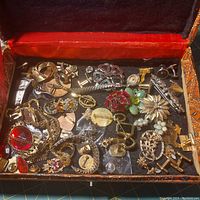 Open jewelry box with assorted vintage jewelry including cufflinks, brooches, pins, and chains scattered inside.