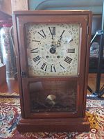 Front view of wooden framed clock with floral and Roman numeral face, showing wear and paint chipping on frame.