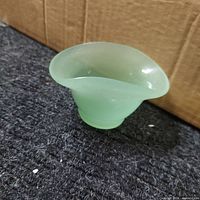 Side angle view of a small light green jadeite glass bowl showing its irregular rounded triangular shape and short pedestal base.