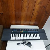 Front view of vintage Casio SA-20 keyboard showing the keyboard, control panel, and power adapter on a white surface against wooden background.