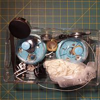Overhead photo showing all items in a clear plastic bin against a cutting mat grid background. Includes watchmaker loupe, tweezers, two blue compartment trays with many small watch parts, small bag of white gaskets, and scattered metal components.