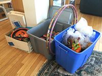 Overview of two plastic bins — grey and blue — filled with books, toys, Halloween decorations, and other items. A box is visible behind containing other objects. Two decorative hula hoops are resting inside the grey bin.
