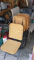 Four wicker folding chairs stacked with woven backs and seats on black metal frames in a storage area.