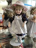 Full view of a porcelain doll wearing a dark floral dress, white eyelet lace apron, and pink lace hat with flowers. Doll stands on a display stand.