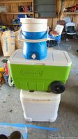 Stacked view of three coolers: top blue and white Igloo serving cooler, middle green Coleman cooler with wheels, bottom white Igloo cooler.