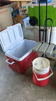 Open red cooler with white lid, green wheeled cooler stacked behind, small red and white Rubbermaid cooler, and folding stool with beige plastic seat and black metal frame.