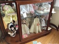 Glass display cabinet with multiple collectible dolls including porcelain dolls in wedding and vintage dresses, mink fur doll, and pastel colored outfits.