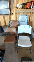 Group of four white and three brown folding chairs stacked and standing, showing plastic and vinyl seats and backs with metal frames in a storage area.