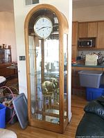 Full view of the grandfather clock inside a room showing oak wood frame, glass front and sides, pendulum and weights inside.