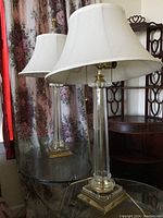 Two matching glass table lamps with white bell-shaped fabric lampshades placed on a glass table near floral curtains and wooden furniture.