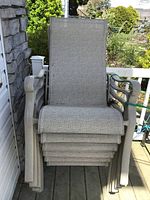 Stack of six weathered patio chairs with beige fabric seating and gray curved arm frames.