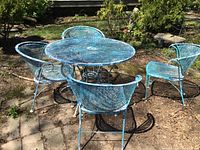 Outdoor photo of round blue metal patio table with mesh top and four matching metal chairs with mesh seating, showing paint peeling and weather wear.