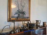 Wide view showing all items arranged on a wooden surface under a framed wall painting, including various silverplate and copper pieces.