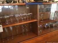 Set of iridescent and clear stemware glasses arranged on wooden shelves inside a glass cabinet.