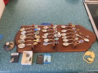 Full view of all collectible spoons arranged on wooden holder, miscellaneous jewelry, thimble, and gold earring on countertop.