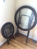 Two round folding tables leaning against corner wall; one large and one small. Glass tops are textured and clear. Dark brown wicker rims and black metal folding legs visible.
