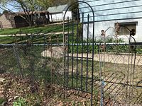 Photo shows a group of metal garden trellises attached to a chain-link fence, four of which have matching vertical bars and decorative fleur-de-lis tops, weathered with rust and some missing parts.