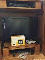 Panasonic VCR on top shelf of wooden media cabinet, Sharp 32in Aquos TV in middle section, Viore DVD player on bottom shelf, Kodak digital frame box and manuals on TV shelf