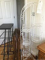 Photo showing the white iron plant stand with decorative scrollwork and multiple round shelves, dark wooden TV trays folded next to it, and the black metal rolling stand behind on hardwood floor.