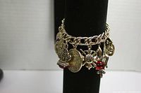 Front view of heavy silver-tone charm bracelet with 9 large charms displayed on a black stand. Charms are detailed and include coins, sunburst motifs, and colored stones.