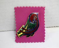 Colorful enameled bird brooch shown on pink fabric background, displaying front view of brooch with intricate swirled pattern and multicolored enamel segments.