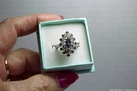 Hand holding the ring in a light blue jewelry box with white cushioned interior, showing ring profile and central blue marquise shaped stone surrounded by clear and dark stones.