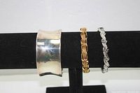 Three bracelets on a black display stand showing front and sides of large silver-tone cuff, gold-tone bracelet with safety chain, and slender silver twisted bracelet.