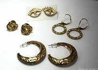 Four pairs of gold-tone costume earrings in a group: large crescent hoop with floral texture, circular wreath rhinestone dangles, love knot studs, and eye-shaped rhinestone studs.