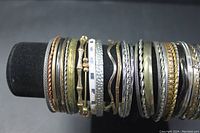Photo showing multiple multicolor bangle bracelets displayed on black velvet tube stand, displaying variety of metal finishes and styles.