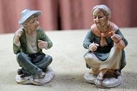 Front and slightly angled view of man and woman porcelain figurines sitting cross-legged on stone bases, showing detailed facial features and clothing.