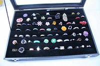 Front view of a large collection of rings organized in rows within a black jewelry display case. Rings feature a variety of designs, sizes, and materials.