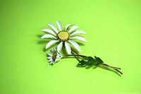 Full view of the vintage large daisy flower brooch on a green background showing white enamel petals, yellow textured flower center, smaller flower bud and green enamel leaf on gold-tone metal stem