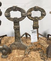 Pair of cast iron andirons positioned upright on wood platform, showing full front view with rustic patina and embedded identification tag.