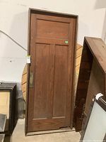 Full view of solid wood swinging door with frame leaning against wall. Shows three-panel door style and attached frame.