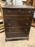 Front view of a dark wood dresser with five drawers and metal handles. The top drawer has a decorative keyhole and ring pulls. Scratches and wear visible on the surface.