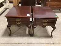 Pair of matching dark wood end tables with cabriole legs, featuring single drawer with ornate brass handles seen from front view on concrete floor.