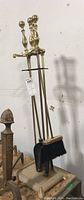 Full view of brass toned fireplace tools including poker, tongs, shovel, and brush held by cast iron base stand.