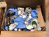 Wide shot of the box containing various mixed china pieces including salt and pepper shakers, a creamer, small plates, and other items all nestled in blue packing material.