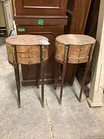 Pair of small vintage boudoir tables shown side by side, wooden round tops with veneer finish missing, each with three front drawers and brass decorative accents on sides and legs.