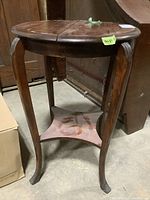 Front angled view of the wooden side table showing curved legs and lower shelf, surface worn and dusty, with green tape label visible.