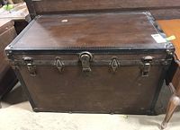 Front view of a large wooden trunk with metal corner protectors, three metal latches on the front, and one broken leather handle on the side.