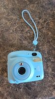 Front view of light blue Fujifilm Instax Mini 11 camera showing lens, flash, wrist strap, and main body.