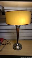 Full view of the lamp on a shelf showing yellow fabric shade, tapered wood and metal base, power cord, and plug.