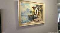 Framed oil painting of a villa by the water with mountains in the background, hung on a wall.