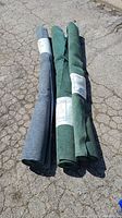 Three rolled outdoor carpets, two green and one blue-grey, laid on cracked asphalt.
