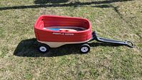 Side view of red and beige plastic Radio Flyer wagon showing body, wheels, and handle on grass.