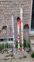 Photo showing 3 pairs of skis and 3 pairs of ski poles leaning against a brick wall outdoors.