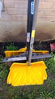 Four yellow snow shovels with metal shafts standing upright against an outdoor wall, showing scoops and handles distinctly.